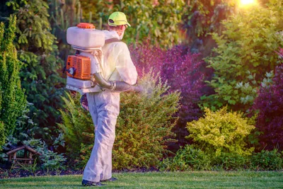 A landscaper in protective gear applying a turf treatment with a backpack mist blower.